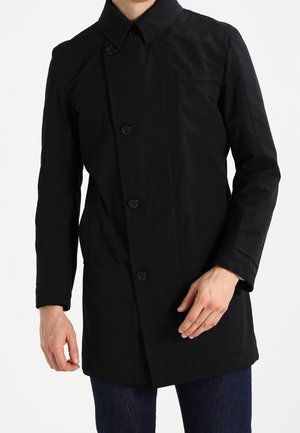 Homme portant un manteau noir boutonné avec un col, visible du cou jusqu'à la mi-cuisse, associé à un jean bleu foncé.