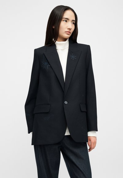 Γυναίκα που φοράει μαύρο φαρδύ σακάκι με διακριτική κεντητή διακόσμηση αστεριών και κυμάτων πάνω από λευκή μπλούζα με ψηλό λαιμό και σκούρο παντελόνι, κοιτάζει προς το πλάι.