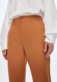 Rostfarbene, taillierte Hosen aus strukturiertem Stoff mit hohem Bund und figurbetonter Silhouette. Kombiniert mit einem weißen, knöpfbaren Hemd.