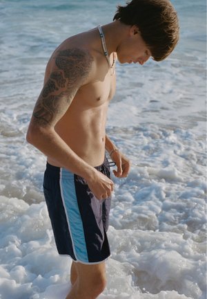 Tânăr cu tatuaj pe braț, purtând pantaloni de baie, stând în apă puțin adâncă din ocean cu valuri și spumă.
