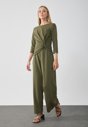 Olivengrønn jumpsuit med vridd midje-design, trekvartlange ermer, vide bukser og svarte stroppehæler.