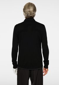 Man med grått hår som bär en svart långärmad tröja och svarta byxor, stående mot en enfärgad vit bakgrund med ryggen mot betraktaren.