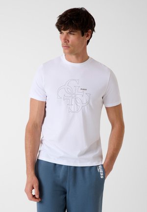 Mann trägt ein weißes Kurzarm-T-Shirt mit einem schwarzen GUESS-Logo im Umriss, kombiniert mit blauen Jogginghosen mit weißem GUESS-Logo.