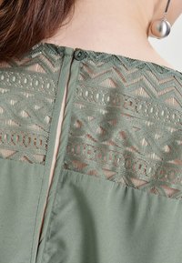 Blusa verde oliva con un respaldo de encaje texturizado con patrones geométricos, un cierre de botón en el cuello y tela suave en la parte inferior.