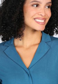 Teal collared garment with a smooth texture; features a v-neckline and subtle stitching details on the front.