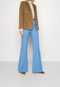 Brun kordfløjl blazer, beige turtleneck, blå klokkebukser og brune spidse hæle, som fremhæver kontrasterende teksturer og farver.