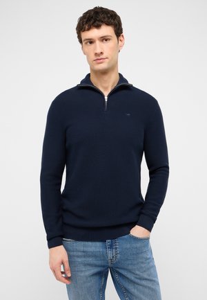 Mann trägt marineblauen Pullover mit Viertelreißverschluss und hellblaue Jeans, steht mit einer Hand in der Tasche vor einfarbigem Hintergrund.