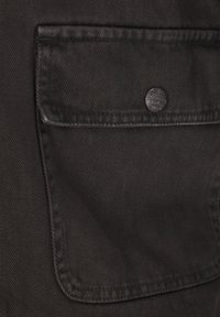 Schwarze strukturierte Stofftasche mit einem Metallknopf mit eingravierter Markenbezeichnung, der auffällig auf dem Kleidungsstück positioniert ist.