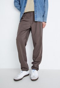 Pantalons marron à coupe décontractée avec une texture lisse, associés à des baskets blanches. La tenue est complétée par une veste en denim et un haut beige.