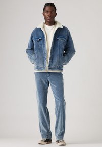 Denimjack met een witte sherpa-kraag, blauwe jeans en zwarte sneakers. De jas heeft voorzakken en een knoopsluiting.