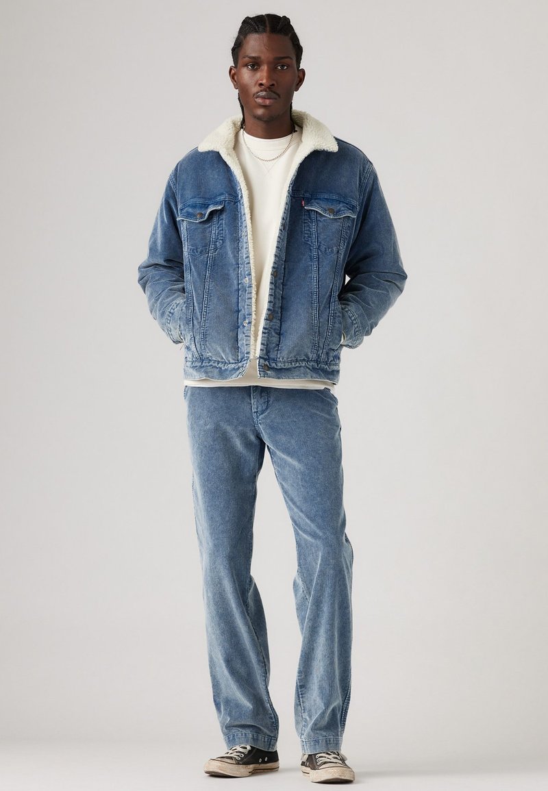 Jeansjacke mit einem weißen Sherpa-Kragen, blauen Jeans und schwarzen Sneakers. Die Jacke hat Fronttaschen und einen Knopfverschluss.