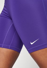 Shorts de sport violet en tissu lisse et extensible, avec des coutures plates et un logo Nike blanc sur le bas à gauche.
