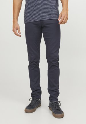 Homme portant un pantalon slim bleu marine foncé et des baskets noires à semelles en caoutchouc, debout devant un fond clair uni.