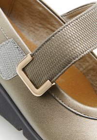 Zapato de bronce metálico con punta redonda, correa elástica texturizada y hebilla en tono dorado. Presenta una textura suave y una suela flexible.