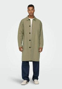 Manteau vert olive présentant une coupe droite, de grandes boutons noirs et un col, associé à un jean bleu foncé et des chaussures blanches.