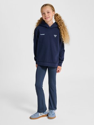 Mädchen mit langen lockigen Haaren steht, trägt einen dunkelblauen Hoodie, passende Hose und hellblaue Sneaker vor einfarbigem Hintergrund.