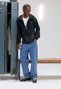 Zwarte jas met rits, wit shirt, blauwe ruime spijkerbroek en zwarte schoenen. Staat vlakbij een grijze kluis met een houten bankje op een neutrale achtergrond.