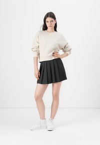Beige fleece genser med senkede skuldre, sammen med et plisserte svart skjørt og hvite sneakers, står mot en ensfarget bakgrunn.