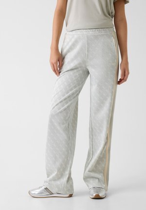 Kobieta ubrana w jasnoszare, wzorzyste spodnie o szerokich nogawkach z beżowymi paskami po bokach oraz srebrne trampki, stojąca na jednolitym tle.