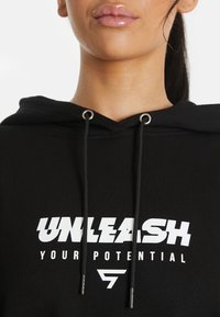 Sudadera negra con un bolsillo canguro en la parte frontal, cordones y "LIBERA TU POTENCIAL" impreso en blanco. Tela suave con un corte relajado.