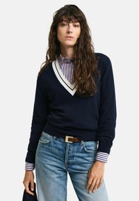 Maglione blu navy con scollatura a V e accenti a righe bianche, abbinato a una camicia a righe. Indossato con jeans azzurri e una cintura marrone.