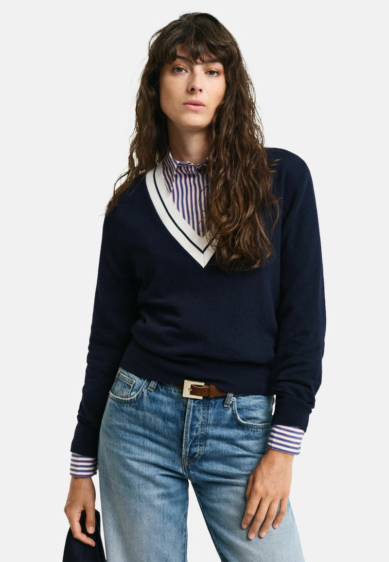 Maglione blu navy con scollatura a V e accenti a righe bianche, abbinato a una camicia a righe. Indossato con jeans azzurri e una cintura marrone.