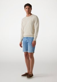Bež pletena dolga majica s dolgimi rokavi, svetlo modri jeans kratke hlače s prevrnjenimi robovi, rjave vezalke, ki stojijo ob enotni ozadju.