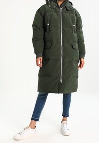 Zielona puchowa kurtka z kapturem, z poziomym pikowaniem, dwoma przednimi kieszeniami i pełnym zamkiem błyskawicznym. Noszona na niebieskich dżinsach i białych tenisówkach.