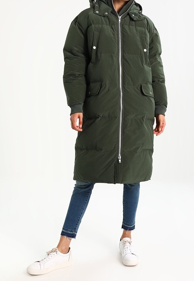 Zielona puchowa kurtka z kapturem, z poziomym pikowaniem, dwoma przednimi kieszeniami i pełnym zamkiem błyskawicznym. Noszona na niebieskich dżinsach i białych tenisówkach.