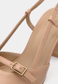 Sandale à talon haut nude avec une tige en cuir lisse, des lanières fines et une boucle en métal doré en accent. Talon épais pour plus de stabilité.