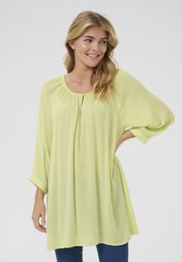 Tunique fluide, légère et transparente de couleur vert clair, avec des manches trois-quarts, un col froncé et un bouton unique sur le devant.