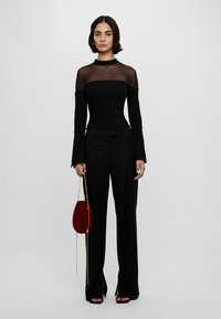 Tenue noire comprenant un haut ajusté à manches longues avec des panneaux transparents et un pantalon large. Un petit sac rouge avec une chaîne dorée est tenu à la main.