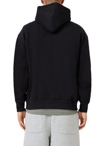 Persona che indossa un felpa nera con il cappuccio alzato, pantaloni tuta grigi con tasche posteriori e una maglietta verde che sbuca da sotto.