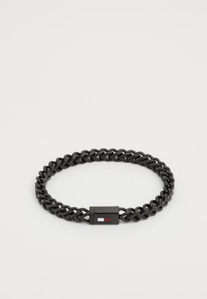 Pulsera negra trenzada hecha de material flexible, con un cierre rectangular que presenta un pequeño acento de logotipo en rojo y blanco.