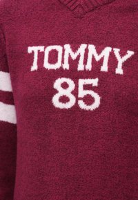 Bordó kötött pulóver V-nyakkal, fehér "TOMMY" és "85" felirattal. Texturált anyag fehér csíkokkal az ujjakon.
