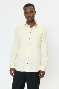 Lichtcrèmekleurig katoenen shirt met lange mouwen en een kraag met knoopsluiting, voorzien van bruine knopen en een losse pasvorm. Zwarte broek zichtbaar eronder.