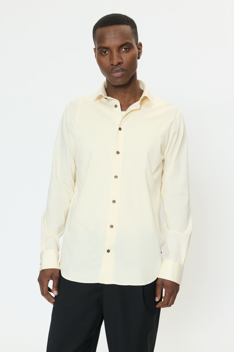 Lichtcrèmekleurig katoenen shirt met lange mouwen en een kraag met knoopsluiting, voorzien van bruine knopen en een losse pasvorm. Zwarte broek zichtbaar eronder.