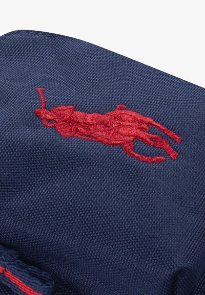 Raudonas siuvinėtas polo žaidėjas ant arklio, ant tekstūruoto tamsiai mėlynos spalvos audinio, greičiausiai drabužio ar aksesuarų dalis.