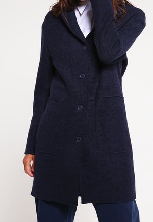 Kobieta ubrana w granatowy rozpinany sweter z dwoma przednimi kieszeniami, na białej koszuli i granatowych spodniach.