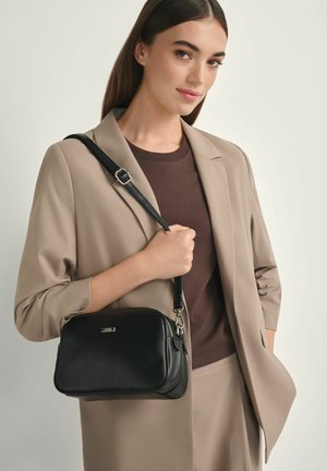 Kobieta ubrana w beżowy garnitur i brązowy top trzyma czarną skórzaną torbę na ramię z srebrną klamrą na jednolitym tle.