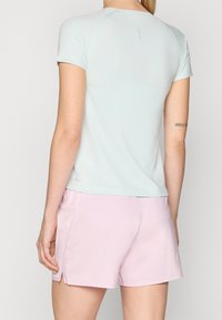 Nainen, jolla on päällään vaaleansininen lyhythihainen paita ja vaaleanpunaiset urheilushortsit, näkyy selkäpuolelta tasaisella taustalla.