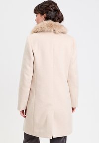 Manteau en laine beige avec un col en fausse fourrure, coupe droite, manches longues et fente au dos. Texture lisse, détails minimalistes.