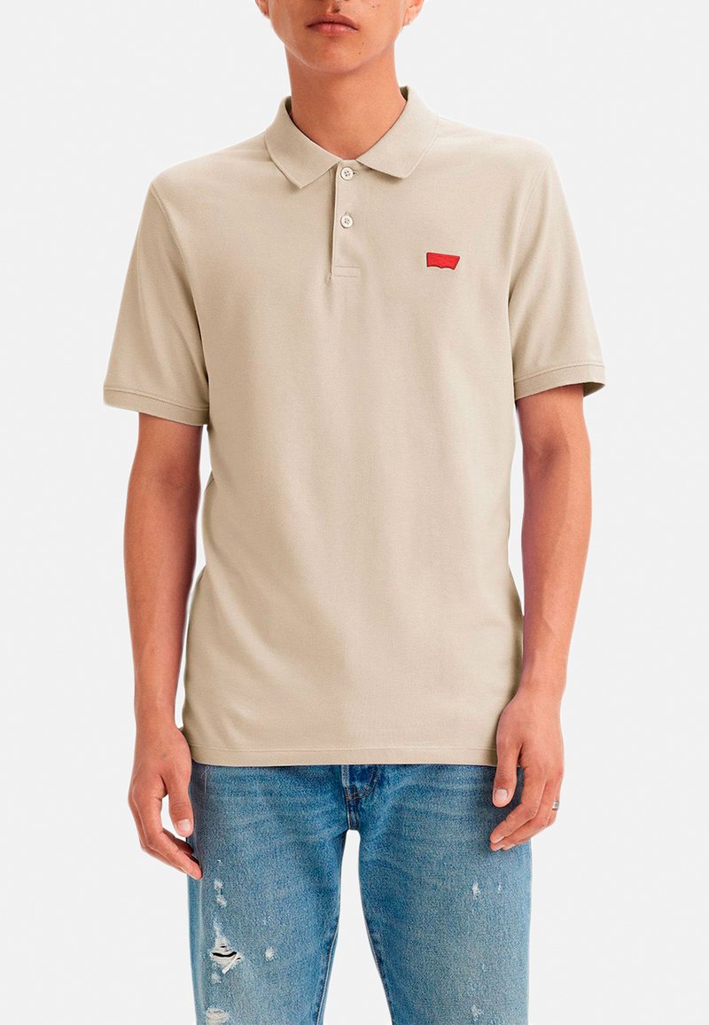 Polo color beige hecho de algodón, con cuello, tres botones y un pequeño logo rojo en el lado izquierdo del pecho. Se lleva con vaqueros azules desgastados.