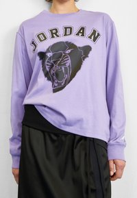 Violet pitkähihainen paita, jossa on graafinen musta-valkoinen pantterisuunnitelma ja "JORDAN"-teksti yläpuolella. Ribattu musta vyötäröyksityiskohta.