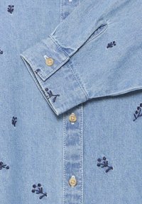 Camicia di jeans blu chiaro con ricami di fiori blu scuro, chiusura con bottoni marroni e maniche arrotolate.