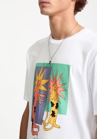 T-shirt en coton blanc arborant un graphique coloré de deux chats avec des éclats d'étoiles, l'un rose et l'autre jaune, sur un fond vert et bleu.