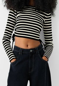 Top corto de rayas negro y blanco con mangas largas y lazos laterales, combinado con pantalones de mezclilla oscuros de cintura alta con costuras visibles.