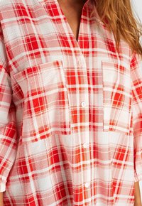 Chemise à carreaux rouge et blanc en tissu léger, dotée d'une fermeture à boutons, de manches courtes et de deux poches poitrine avec une texture douce.