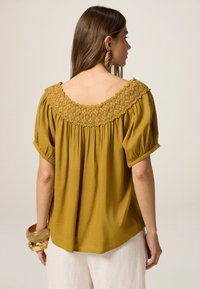 Mosterdgele blouse met een brede, prachtig geborduurde halslijn, korte puffmouwen en een losse pasvorm, gecombineerd met een gouden armband om de pols.