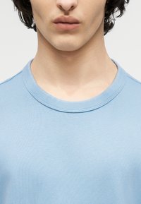 Νεαρός που φοράει ανοιχτό μπλε μπλουζάκι με στρογγυλή λαιμόκοψη, με ορατό λαιμό και κάτω μέρος προσώπου σε απλό φόντο.